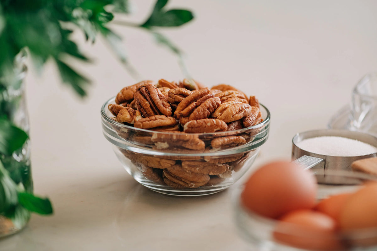 Glass bowl of pecans on a kitchen counter with eggs and a measuring cup in the foreground.