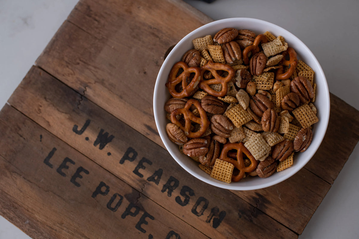 Assorted snack mix in a white bowl on a wooden surface with engraved text.