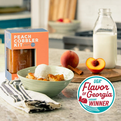 Peach Cobbler Kit box with peach cobbler in bowl topped with ice cream.  Sliced peach, cinnamon sticks and bottle of milk on wooden board.  Flavor of Georgia winner logo