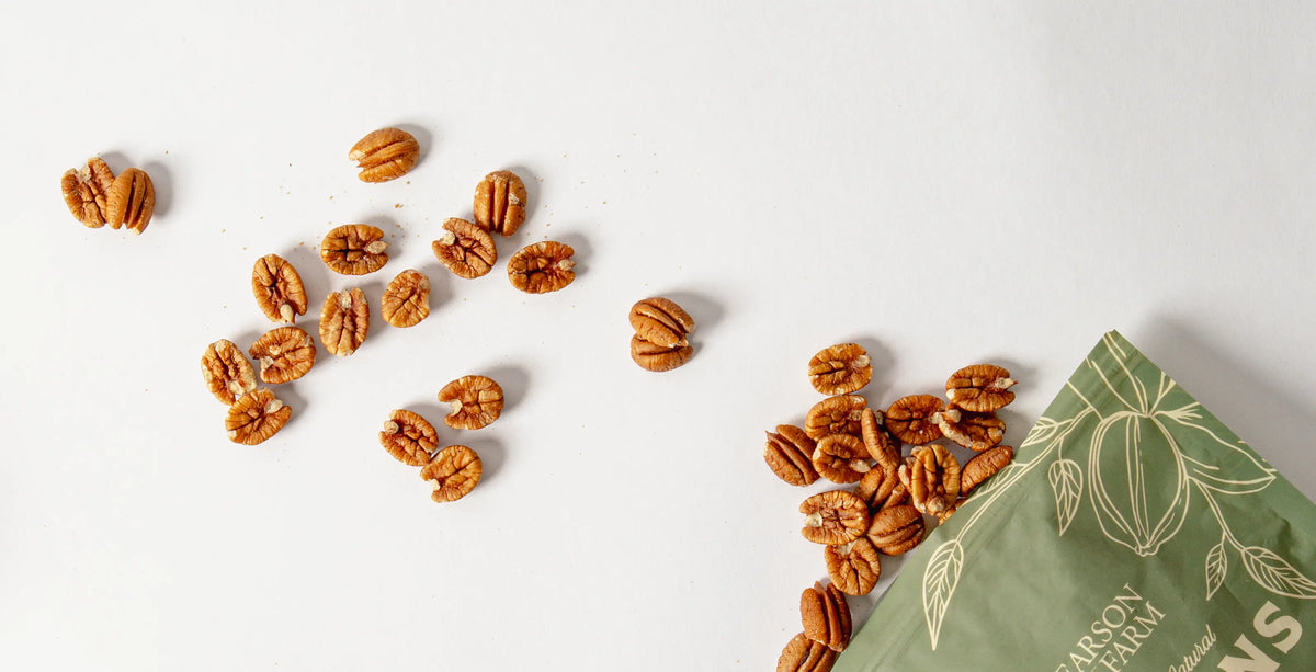 Pecans scattered on a white surface with a green bag featuring pecan design.