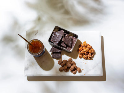 Marble cutting board with a tin of dark chocolate bark, scattered pecans and jar of jelly